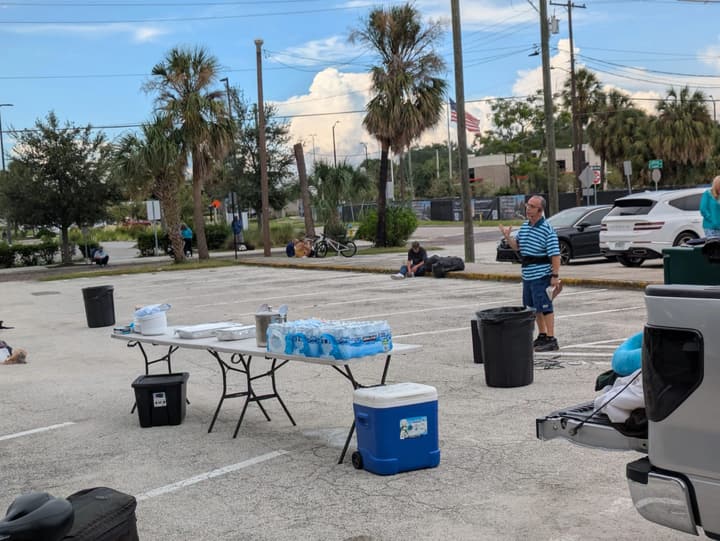 A volunteer speaks beside a folding table with cases of bottled water, a cooler and supplies in a palm-lined parking lot