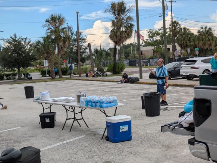 Folding table set with bottled water and serving supplies, a blue cooler and trash cans ready before the meal