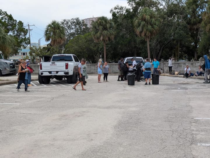 People gather near a pickup truck in a tree-lined parking lot during the Tuesday meal