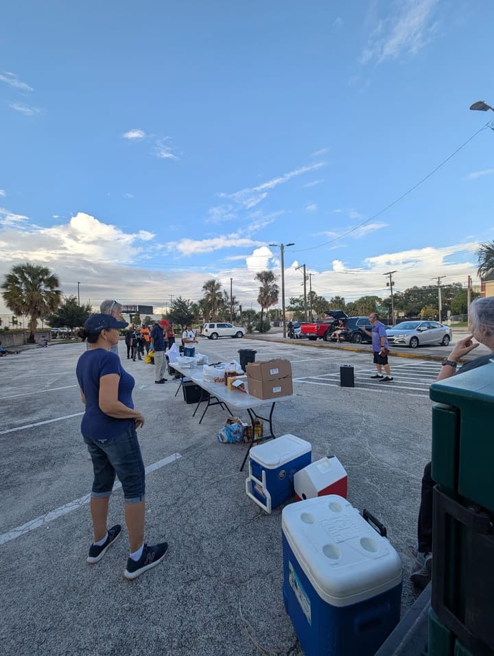 Volunteers set up folding tables with boxes of food and coolers in a parking lot under a wide blue sky