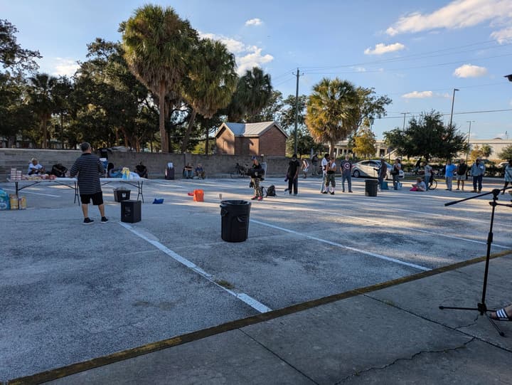 People line up across a sunny parking lot toward the food table, with a microphone stand set up for music