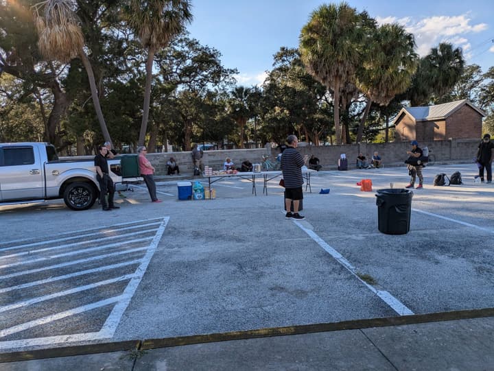 Late-afternoon gathering in a palm-shaded parking lot, with a food table, coolers and people waiting