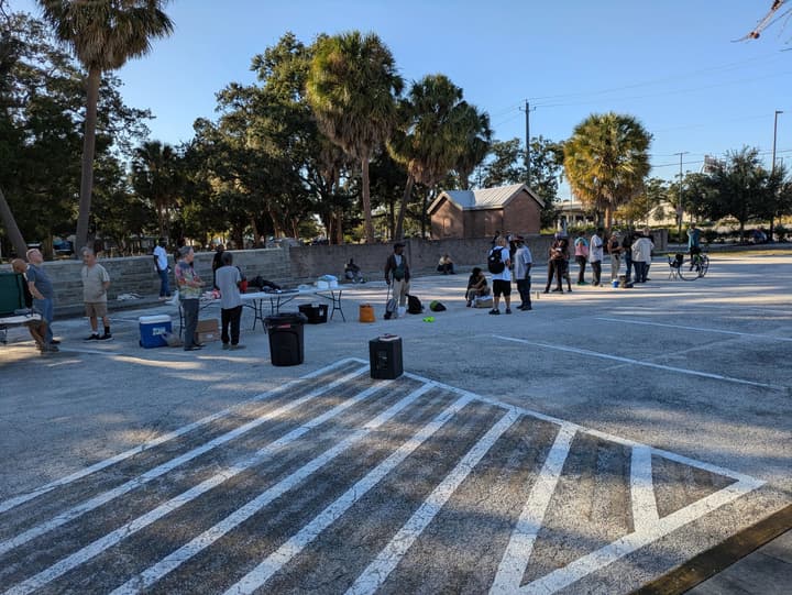 People gather around a food table and a portable speaker in a sunlit parking lot lined with palm trees