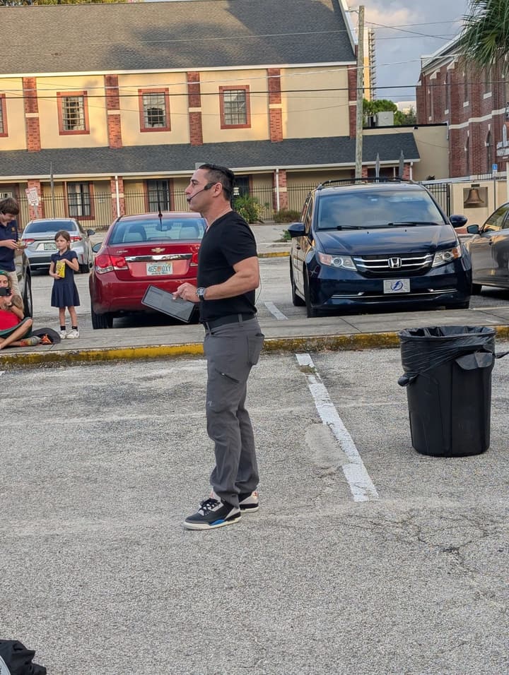 A man with a headset microphone speaks to the gathering in the parking lot