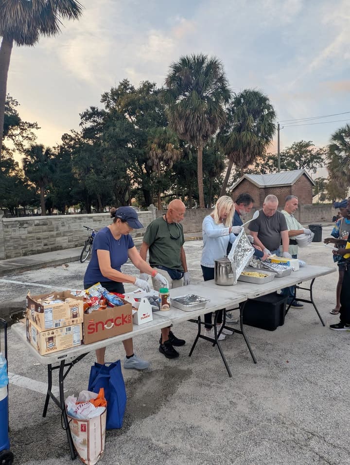 Gloved volunteers serve hot food from foil pans along a row of folding tables at dusk