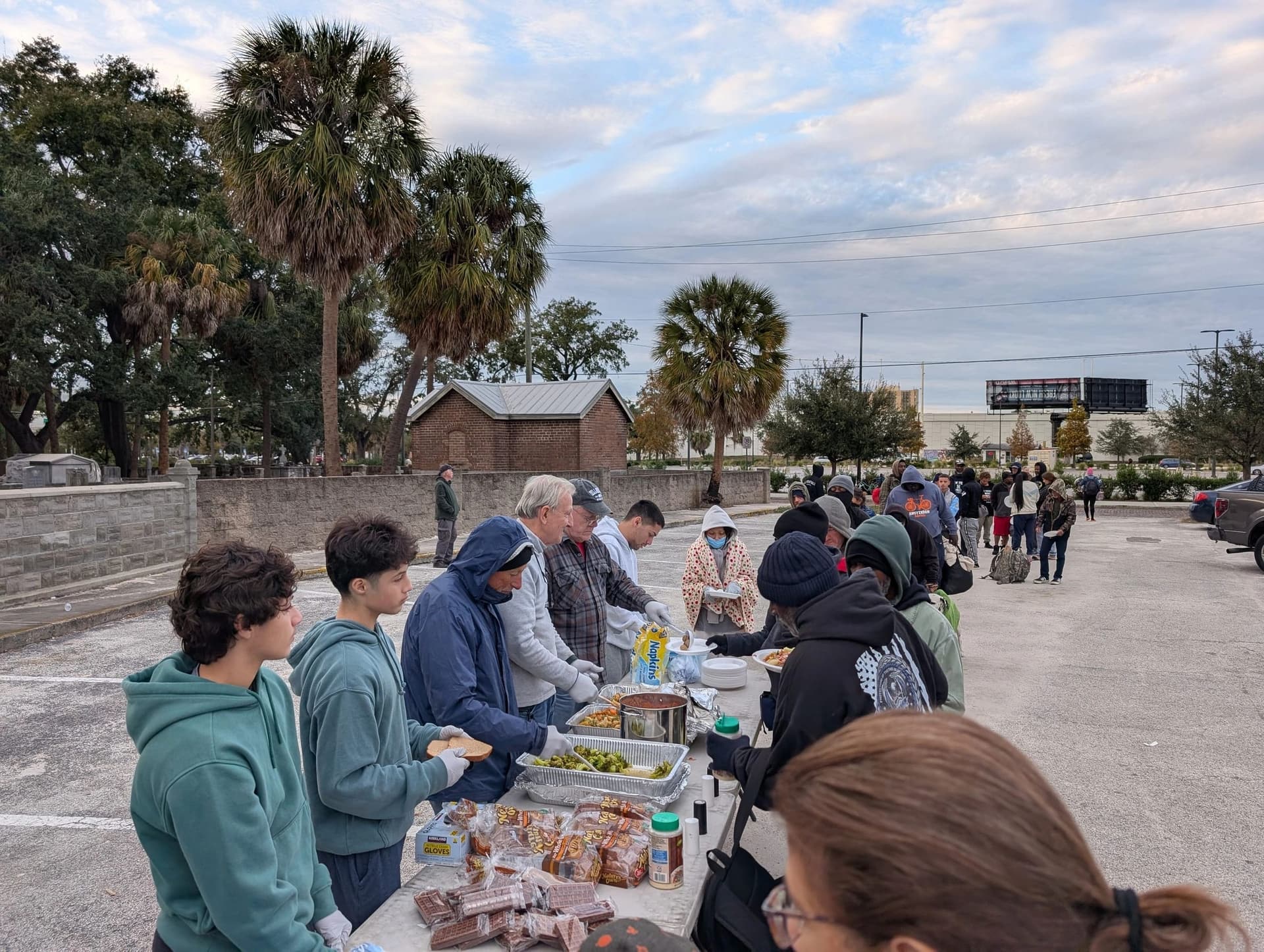 Volunteers of all ages serve hot food, snacks and water to a long line of people in a palm-lined parking lot