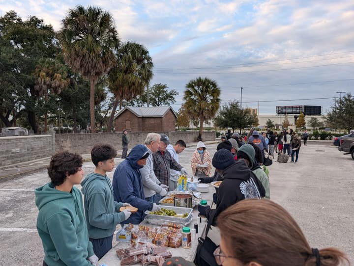 Volunteers of all ages serve hot food, snacks and water to a long line of people in a palm-lined parking lot