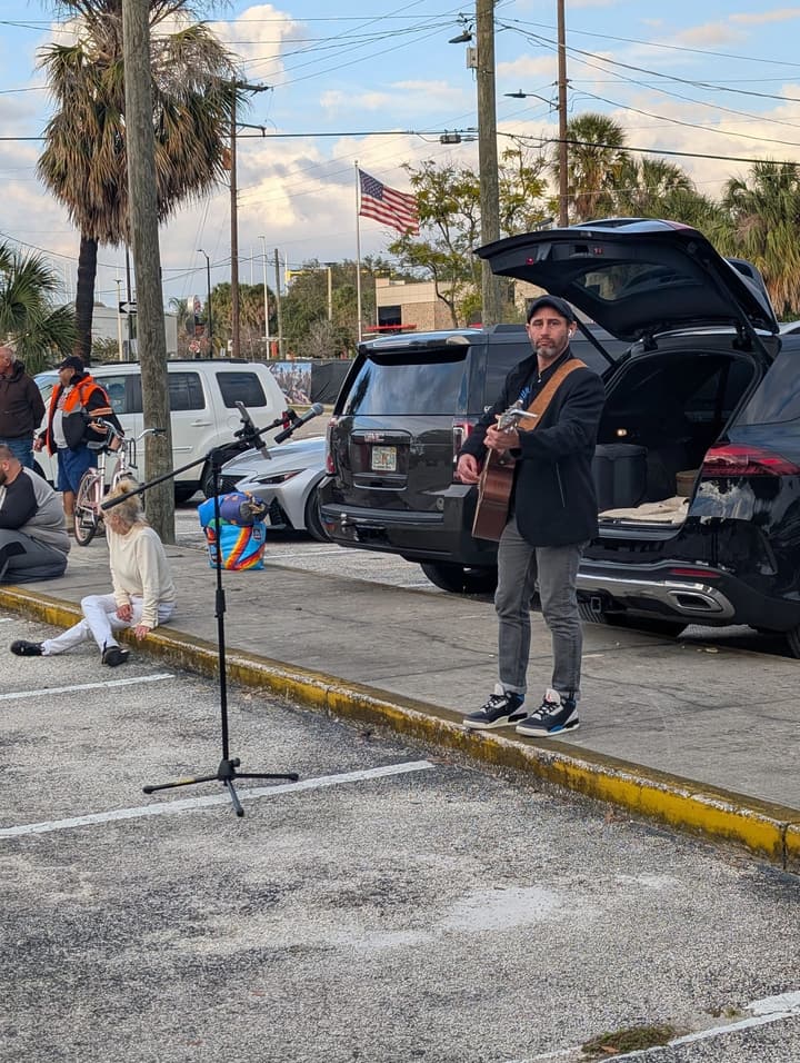 A musician with an acoustic guitar plays at a microphone stand beside the parking lot