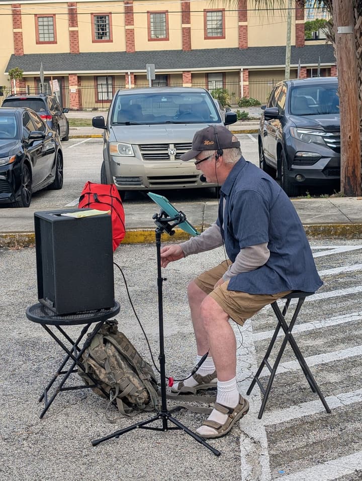 A seated musician with a headset and tablet plays music through a speaker in the parking lot