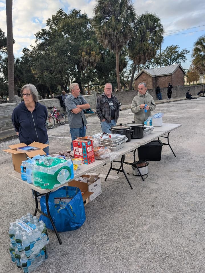 Volunteers stand behind folding tables set with slow cookers, foil pans, bread and bottled water