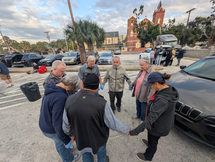Volunteers join hands in a prayer circle in the parking lot, with a red-brick church in the background