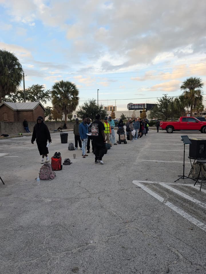 A long line forms across the parking lot at dusk, with a music stand and speaker set up nearby