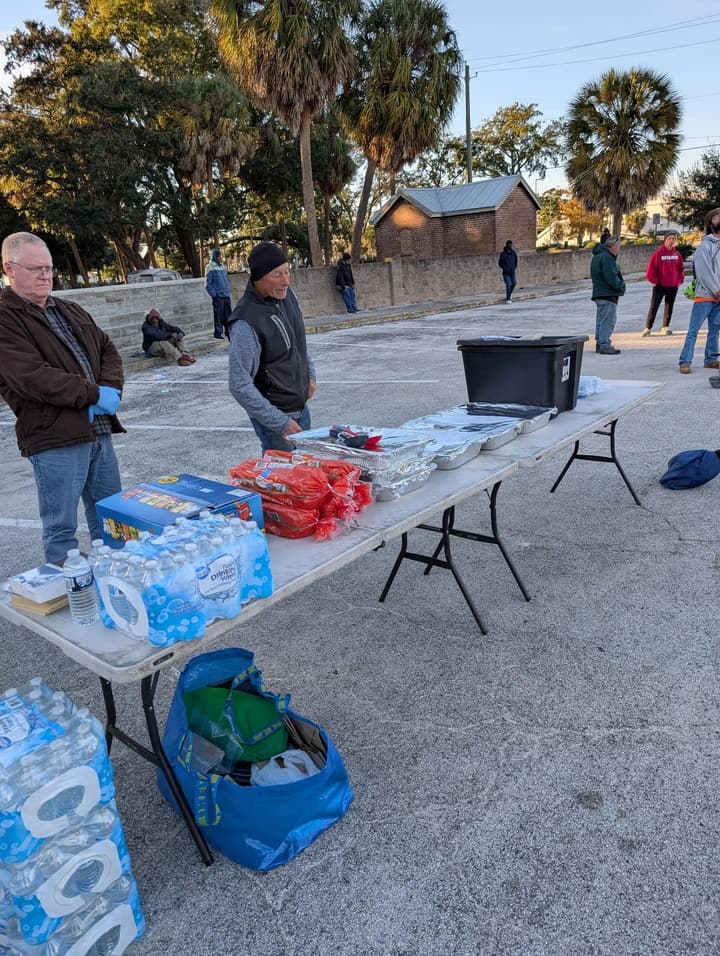 Volunteers in gloves wait behind a table of foil-covered pans, chips and cases of bottled water