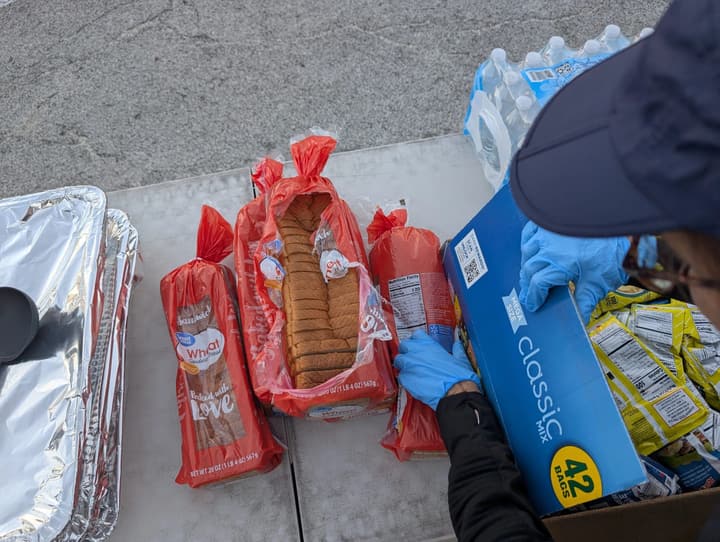 A gloved volunteer unpacks loaves of bread and a box of chips next to pans of hot food