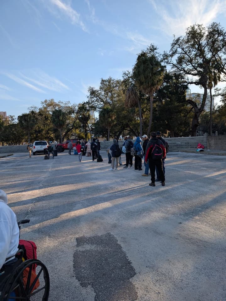 People gather in the sunlit parking lot under oak and palm trees as the meal begins