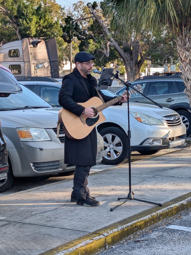 A guitarist in a dark coat and flat cap sings at a microphone stand during the meal