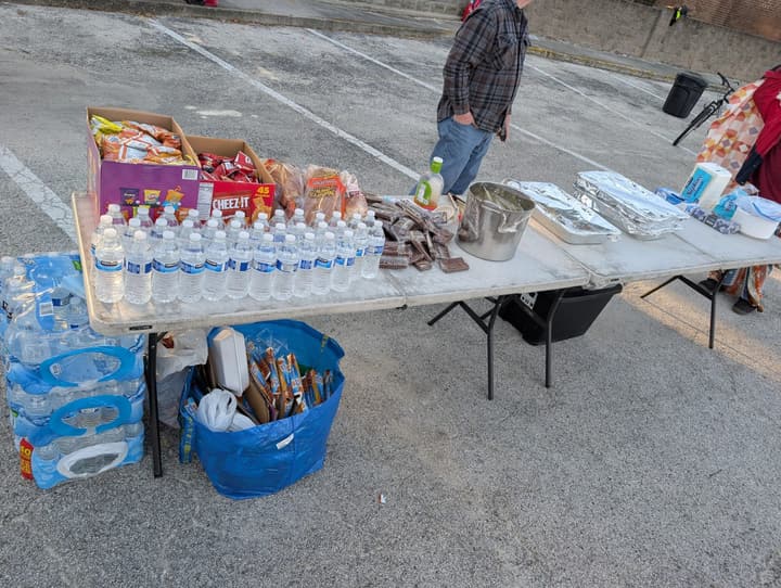Folding tables set with rows of bottled water, snacks, brownies, a stockpot and foil pans of hot food