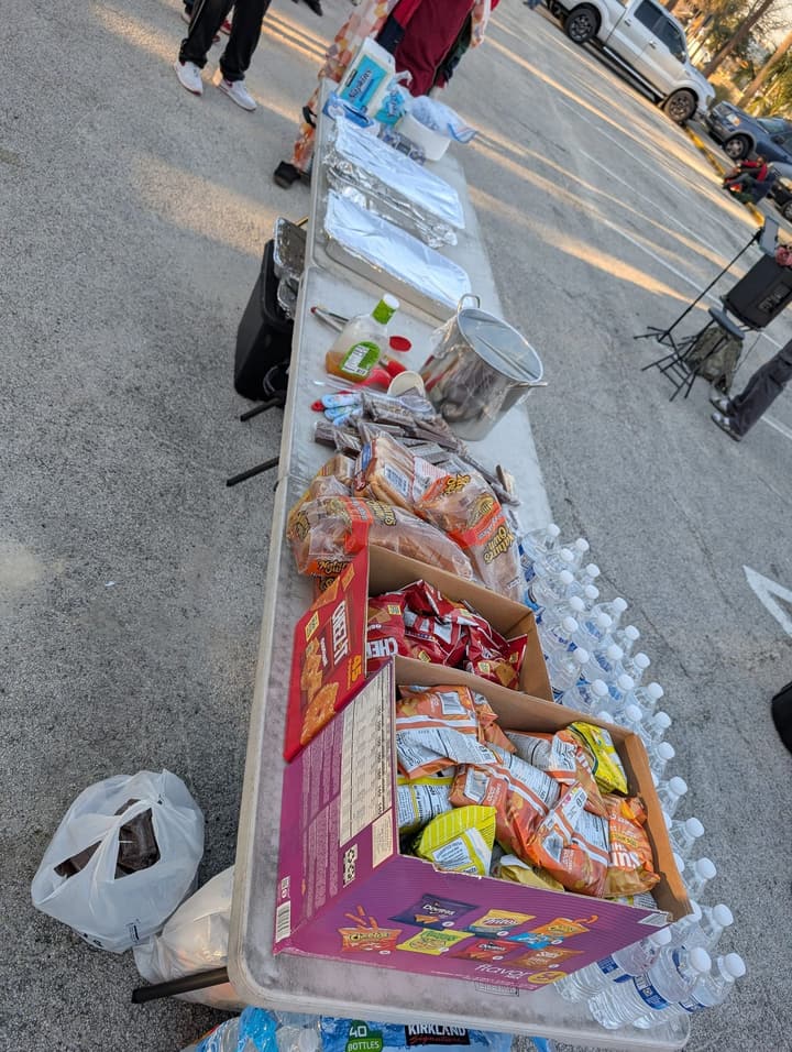 A long serving table lined with bottled water, chips and pans of hot food, with music set up nearby