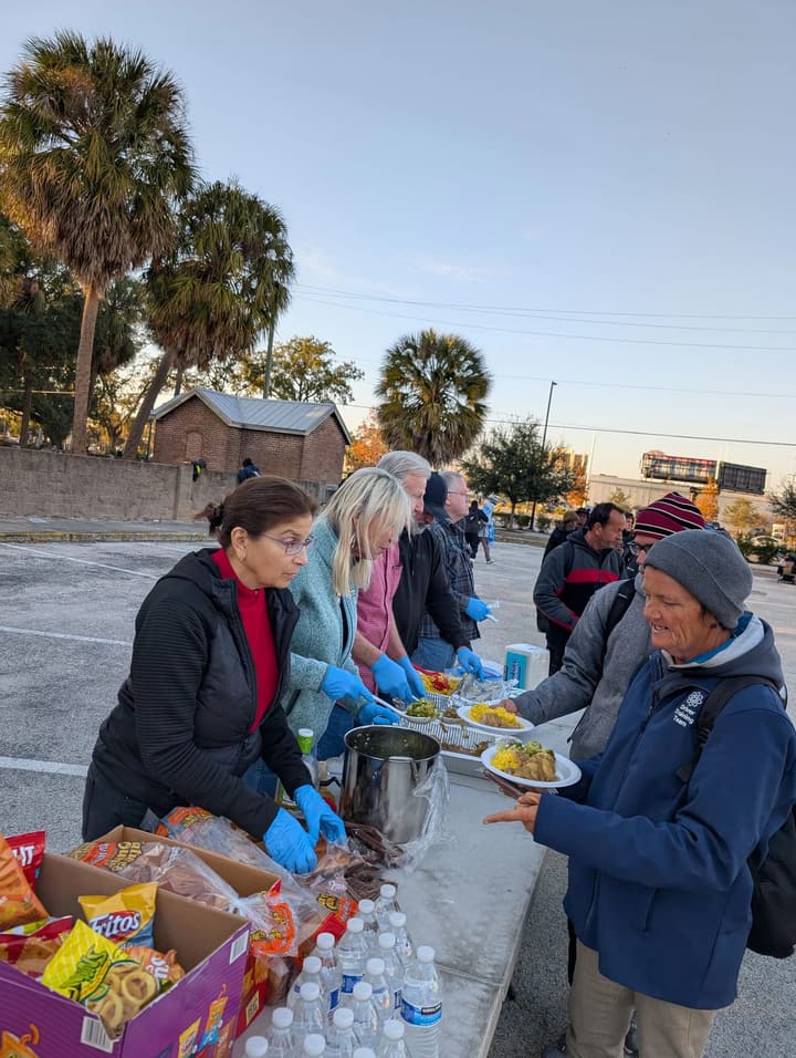 Gloved volunteers hand a plate of hot food to a smiling person in a knit cap at the serving line