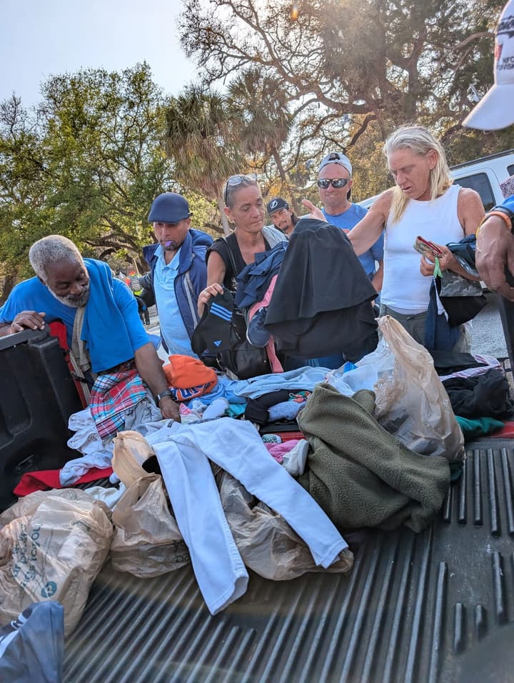 People gather around a pickup truck bed, choosing donated clothing together in the afternoon sun