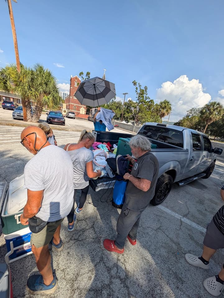 Volunteers sort donated clothing at a pickup truck, with a red-brick church steeple behind them
