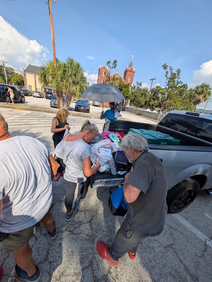 A volunteer holds an umbrella for shade as others sort donated clothing at a pickup truck