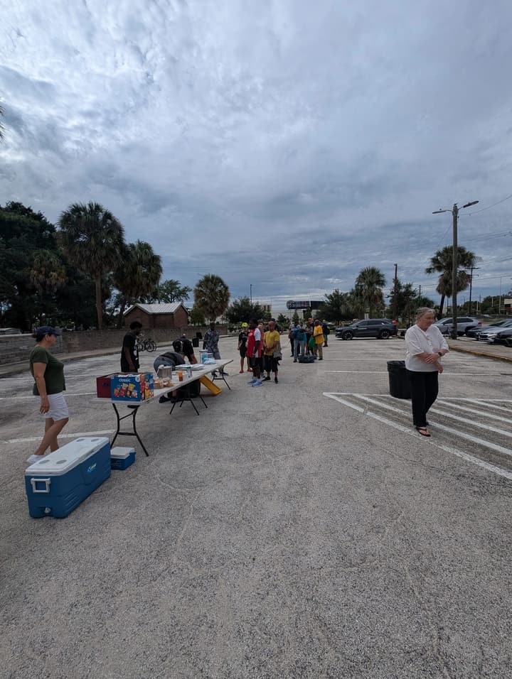 Volunteers set up a serving table and coolers as people begin to gather under a cloudy sky